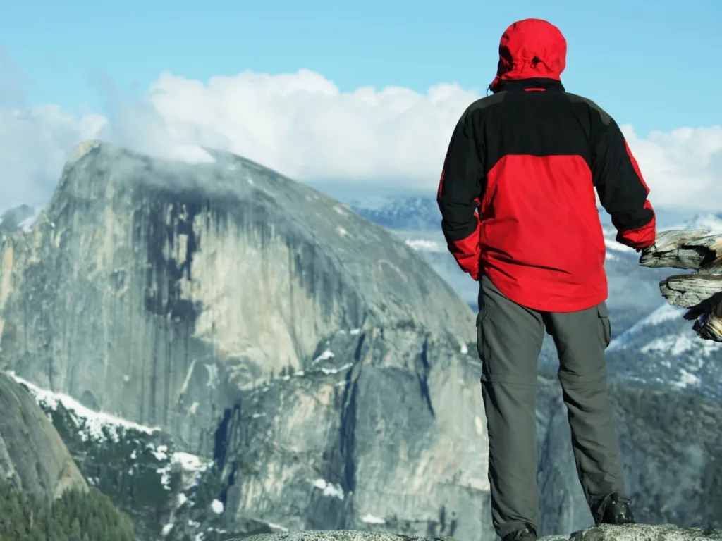 Man in red jacket 
 looking at a vista of Half Dome in Yosemite National Park.
